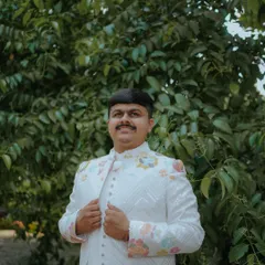 A classic portrait of the groom, looking dapper in his floral-embroidered bandhgala jacket against a natural green background.
