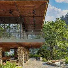 The covered terrace of a building at Taj Rishikesh, featuring stone pillars and a wooden ceiling that frame views of the surrounding greenery. This space exemplifies our use of breathable corridors and passive design to create comfortable, climate-responsive areas.