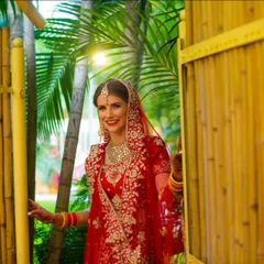 A beautiful bride in a traditional red lehenga, making her entrance through a rustic bamboo gateway. Her smile radiates happiness and anticipation for the ceremony ahead.