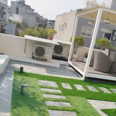 This terrace design combines a stone and grass pathway with a raised, covered seating area under a white pergola, creating different zones for relaxing.