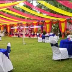 An outdoor wedding reception setup on a lawn with a canopy of yellow and pink drapes and guest tables dressed in blue and white.