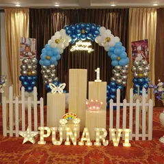 A blue and silver balloon ring arch set up in a party hall. This decoration includes a white picket fence, marquee letters, and pedestals for a clean and classic birthday look.