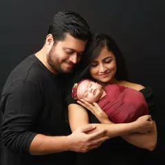 A classic family portrait against a simple black background. The new parents are smiling as they gaze down at their sleeping newborn, who is swaddled in a rich maroon wrap with a matching floral headband.
