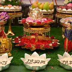 A close-up of a naming ceremony decor piece. The two-tiered brass lotus stand holds a Ganesha idol, flowers, and fruits for the pooja.
