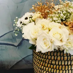 A rustic-chic floral arrangement in a woven basket. This bouquet combines white roses with delicate baby's breath and dried elements, offering a soft and textured look for any room.