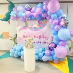 A magical mermaid theme birthday decoration using pastel blue and purple balloons. The setup includes a mermaid tail balloon and a themed backdrop for the cake table.