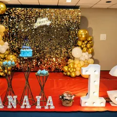 A glamorous 13th birthday party setup with a gold shimmer wall, a 'Happy Birthday' neon sign, and large marquee numbers. The gold and white balloons complete the luxurious look.