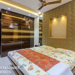 A bedroom in a Vashi row house designed with a rich, golden-upholstered headboard that includes a lit niche. The glossy, wood-grain wardrobe with a custom gold-patterned band adds a touch of opulence and warmth.