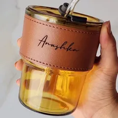 A close-up of the personalized glass sipper with a leather sleeve. The name is elegantly engraved on the sleeve, and the amber-colored glass gives it a premium look.