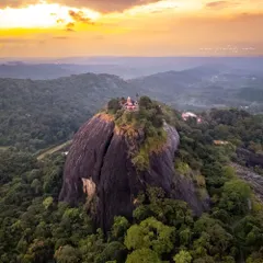 An aerial shot of an ancient Shiva temple perched atop a massive rock formation, surrounded by the lush forests of South Canara. The setting sun casts a warm glow on this historic site mentioned in the Ramayana.