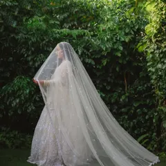 A mysterious and beautiful shot of the bride enveloped in her long, delicate veil against a lush green backdrop.