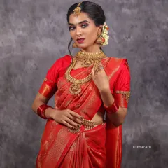 A classic studio portrait showcasing a traditional bridal look. The controlled lighting highlights the rich color of the red saree and the intricate details of the temple jewelry.