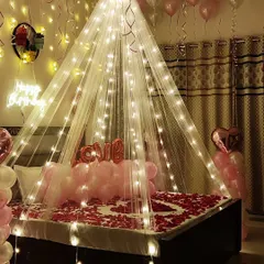 To create an unforgettable romantic birthday surprise, I design complete room decorations. This setup features a canopy draped with fairy lights over the bed, a floor and bed covered in rose petals, and pink balloons to create a warm and intimate atmosphere.