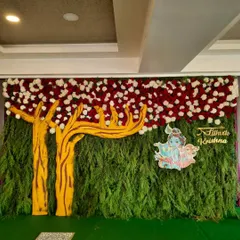 A close-up of the Krishna-themed backdrop. The design features a tree cutout against a wall of green foliage, with a custom nameplate and a colorful depiction of Krishna, perfect for a child's naming ceremony.