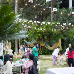 A wide shot of the musical garden party in full swing, with guests mingling, kids playing in the ball pit, and activity tables set up for creative fun under a canopy of string lights.