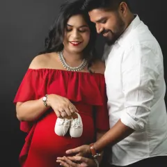 A classic maternity pose with a prop that says it all. The couple holds a pair of tiny baby shoes, symbolizing the new life they are waiting for, captured against a dramatic black backdrop.