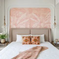 A serene and stylish bedroom featuring a custom upholstered bed set against a soft pink, palm-leaf patterned accent wall. The design is completed with elegant pendant lights and carefully chosen textiles to create a tranquil retreat.