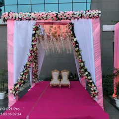 A beautiful stage or mandap setup in a pink and white theme. The entrance to the stage is framed with floral garlands, and two ornate chairs are placed for the couple.