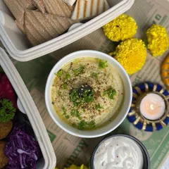 A closer look at our creamy hummus, garnished with parsley and sesame seeds, served with a side of dip. It's the star of our Pita Hummus platter.