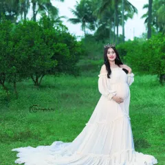 A portrait of pure elegance. The mother-to-be looks regal in a flowing white gown and a delicate crown, set against the natural green backdrop of our exclusive outdoor location.