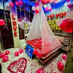 This 'Just Married' room decor is all about celebration. The room is filled with red, pink, and white balloons, with a floor covered in petals, a heart shape on the floor, and a canopy over the decorated bed.