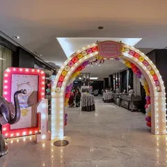 A grand carnival entrance archway for an indoor event. The arch is decorated with colorful balloons and marquee lights, flanked by light-up signs and a friendly elephant prop to welcome guests to the celebration.