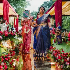 A bride's journey is a cherished family affair. Here, she walks down the golden aisle, accompanied by relatives performing a timeless ritual, all while surrounded by vibrant red floral decor that symbolizes love and celebration.