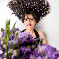 A creative beauty shot from the 'WILD LAVENDER' series. The composition uses a foreground of flowers to frame the model, whose hair is adorned with lavender, creating a whimsical and artistic portrait.