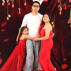 A classic family pose against a romantic red backdrop with hanging roses. The daughter hugs her father while the expecting mother looks on, creating a warm and connected family portrait.