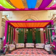 A lively Mehendi stage setup with a colorful canopy of orange, pink, and green drapes, complete with comfortable seating and rustic hanging planters.