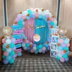 A beautiful gender reveal party setup with a full balloon ring in pastel pink, blue, and purple. The decor includes 'BABY' blocks, themed foil balloons, and a shimmer curtain backdrop.