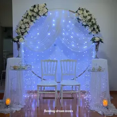 An elegant and simple anniversary backdrop. This setup features a circular frame draped with white fabric, fairy lights, and white floral arrangements, creating a perfect photo opportunity for the happy couple.