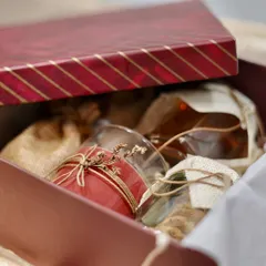 A peek inside a rustic-themed gift box being prepared. This hamper includes a scented candle, jars of gourmet food, and items in burlap sacks, all packed into a deep red box.