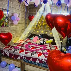 A romantic birthday surprise with a canopy over the bed, lit with fairy lights. The room is filled with purple and pink balloons, large red heart foil balloons, and a heart made of rose petals on the bed.