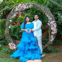A lovely couple portrait in front of a rustic floral wreath. The mom-to-be's vibrant blue gown stands out beautifully against the garden backdrop, and they are making a heart shape with their hands on the baby bump.