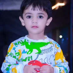 A close-up portrait of a young boy in a colorful dinosaur-themed shirt, looking directly at the camera.