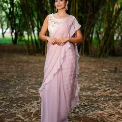 This lavender pre-draped saree features delicate ruffles and sequin work. It's paired with a sleeveless white blouse with intricate gold and pearl embroidery on the neckline.
