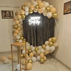 A chic and modern birthday backdrop with a circular frame, a garland of gold, cream, and white balloons, and a 'Happy Birthday' neon sign against a black shimmer curtain.