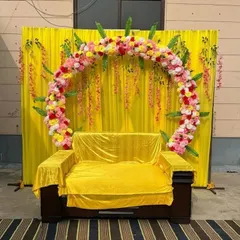 For this Haldi ceremony, I used a simple yellow drape backdrop and added a semi-circular arch of pink and yellow artificial flowers. The setup provides a beautiful and vibrant frame for the seating area.