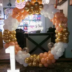 A classic balloon ring in gold, white, and peach for a first birthday. This decoration includes a neon sign and a light-up number '1', creating a warm and inviting photo corner.