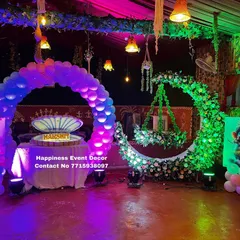 A vibrant naming ceremony setup with a mix of traditional and modern elements. It includes a green floral moon-gate with a hanging cradle, a balloon arch, and colorful lighting to brighten up the venue.