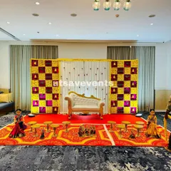 This baby shower setup features a beautiful floral checkered backdrop in yellow and magenta. The white and gold divan provides elegant seating, with traditional dolls and urlis completing the stage.