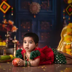 A classic portrait from a traditional photoshoot. This little one, dressed in a gorgeous red and green outfit, is captured in a moment of quiet curiosity amidst a setting filled with cultural details.