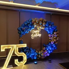 A grand 75th birthday celebration with a blue and gold theme. A full ring of metallic balloons frames a "Happy Birthday" neon sign, complemented by large light-up numbers for the milestone.