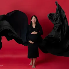 A powerful solo maternity portrait featuring a woman in a black gown against a vibrant red background. The gown's fabric is tossed to create a dynamic, wing-like shape, resulting in a bold and modern studio photograph.