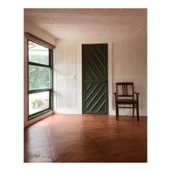 A simple, uncluttered room in the farmhouse featuring a diagonally panelled green door and a single wooden chair. The design emphasizes minimalism and the use of natural light from the large window to create a tranquil space.