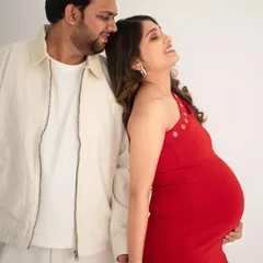 A candid and joyful portrait of an expecting couple. The mom-to-be, in a vibrant red dress, leans back against her partner with a happy smile, basking in the moment.