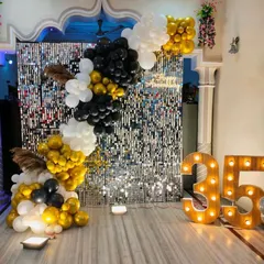 A sophisticated backdrop for a 35th birthday, featuring a silver shimmer wall that adds texture and shine. The asymmetrical balloon garland in gold, black, and white provides a modern and elegant frame, complemented by large light-up marquee numbers.