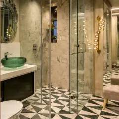 This master bathroom features a stunning geometric floor made of green and white marble, a floating vanity with a green glass basin, and a large Venetian mirror. The glass shower enclosure keeps the space feeling open and bright.