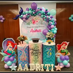 A beautiful Little Mermaid setup for Aadriti's 7th birthday, featuring a rich color palette of purple and teal. The decor includes a mermaid tail balloon sculpture and character props.